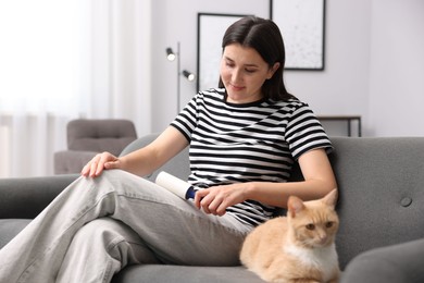 Photo of Woman with lint roller removing pet's hair from clothes and her cat on sofa at home