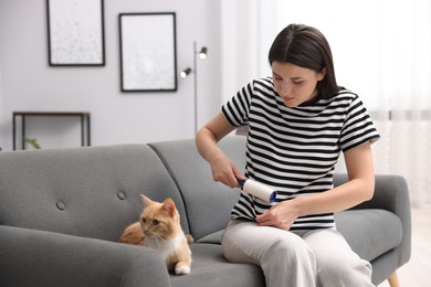 Photo of Woman with lint roller removing pet's hair from clothes and her cat on sofa at home