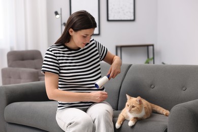 Photo of Woman with lint roller removing pet's hair from clothes and her cat on sofa at home