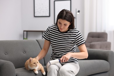 Photo of Woman with lint roller removing pet's hair from clothes and stroking her cat on sofa at home