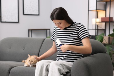 Photo of Woman with lint roller removing pet's hair from clothes and stroking her cat on sofa at home