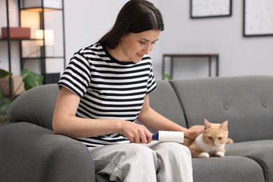 Photo of Woman with lint roller removing pet's hair from clothes and stroking her cat on sofa at home