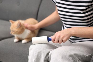 Photo of Woman with lint roller removing pet's hair from pants and stroking her cat on sofa, closeup