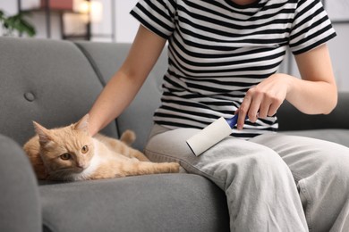 Photo of Woman with lint roller removing pet's hair from pants and stroking her cat on sofa at home, closeup