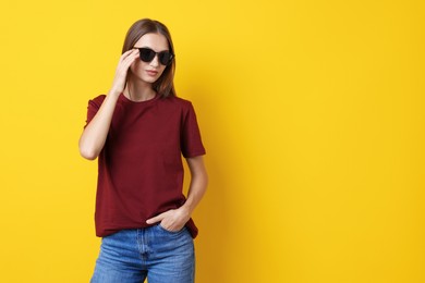 Photo of Woman wearing stylish burgundy t-shirt and sunglasses on yellow background. Space for text