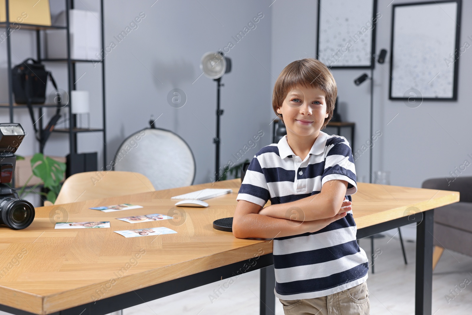 Photo of Choice of profession. Little boy pretending to be photographer leaning on wooden table with camera and pictures in studio