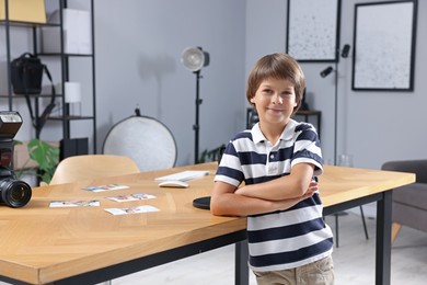 Photo of Choice of profession. Little boy pretending to be photographer leaning on wooden table with camera and pictures in studio