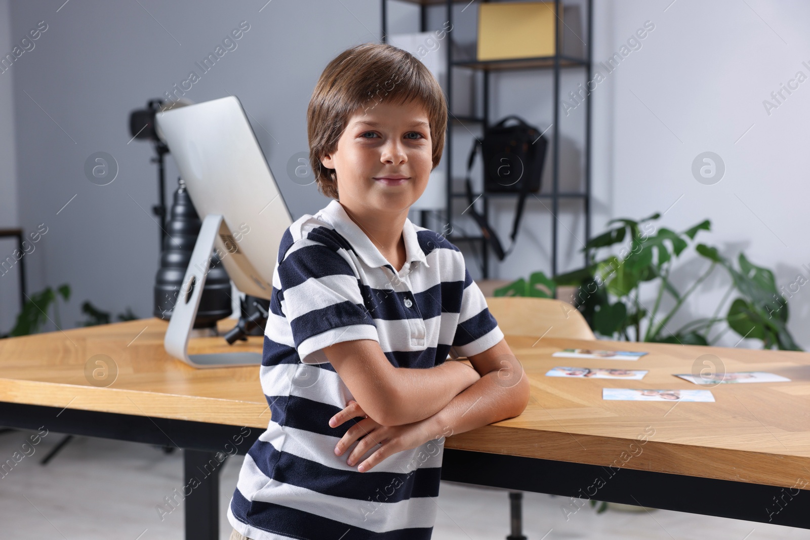 Photo of Choice of profession. Little boy pretending to be photographer leaning on wooden table with computer and pictures in studio