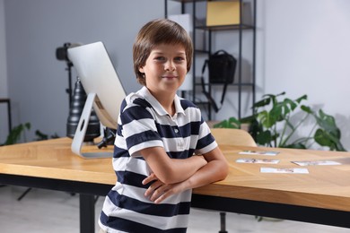 Photo of Choice of profession. Little boy pretending to be photographer leaning on wooden table with computer and pictures in studio