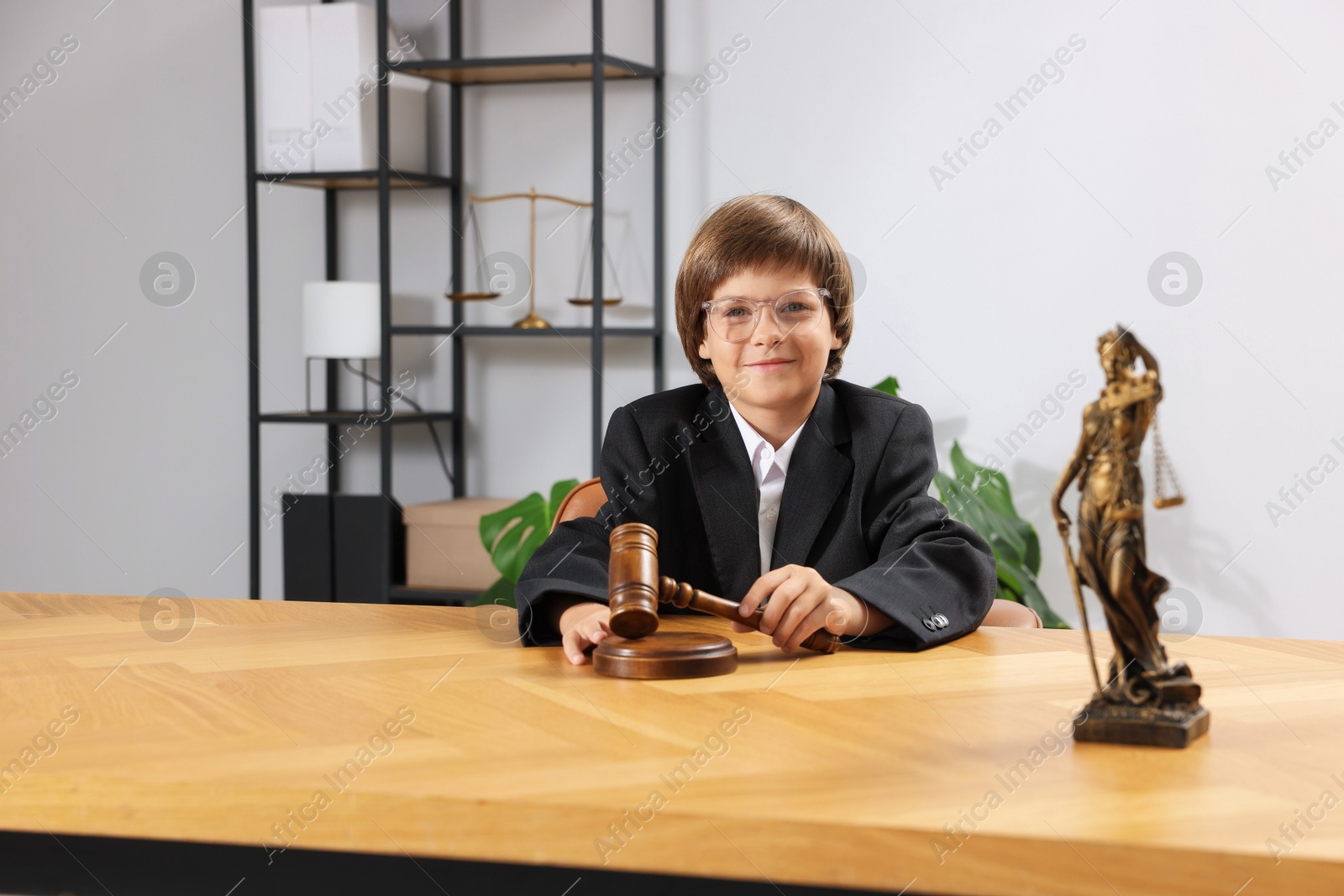 Photo of Choice of profession. Little boy in glasses with judge's gavel and statue of Lady Justice pretending to be lawyer at wooden table in office
