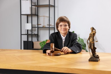 Photo of Choice of profession. Little boy in glasses with judge's gavel and statue of Lady Justice pretending to be lawyer at wooden table in office