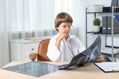 Photo of Choice of profession. Little boy in glasses and uniform with lungs x-ray pretending to be doctor at table in clinic