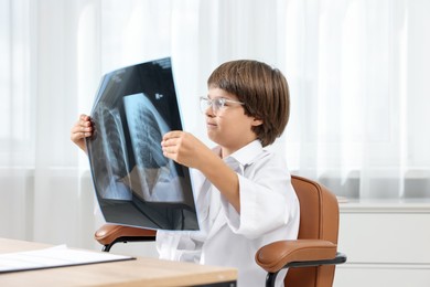 Photo of Choice of profession. Little boy in glasses and uniform with lungs x-ray pretending to be doctor at table in clinic
