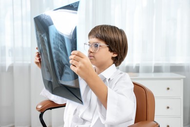 Photo of Choice of profession. Little boy in glasses and uniform with lungs x-ray pretending to be doctor on chair in clinic