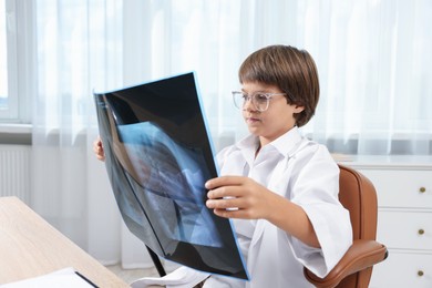 Photo of Choice of profession. Little boy in glasses and uniform with lungs x-ray pretending to be doctor at table in clinic