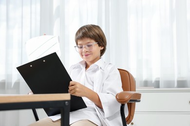 Photo of Choice of profession. Little boy in glasses and uniform with patient's medical card pretending to be doctor on chair in clinic