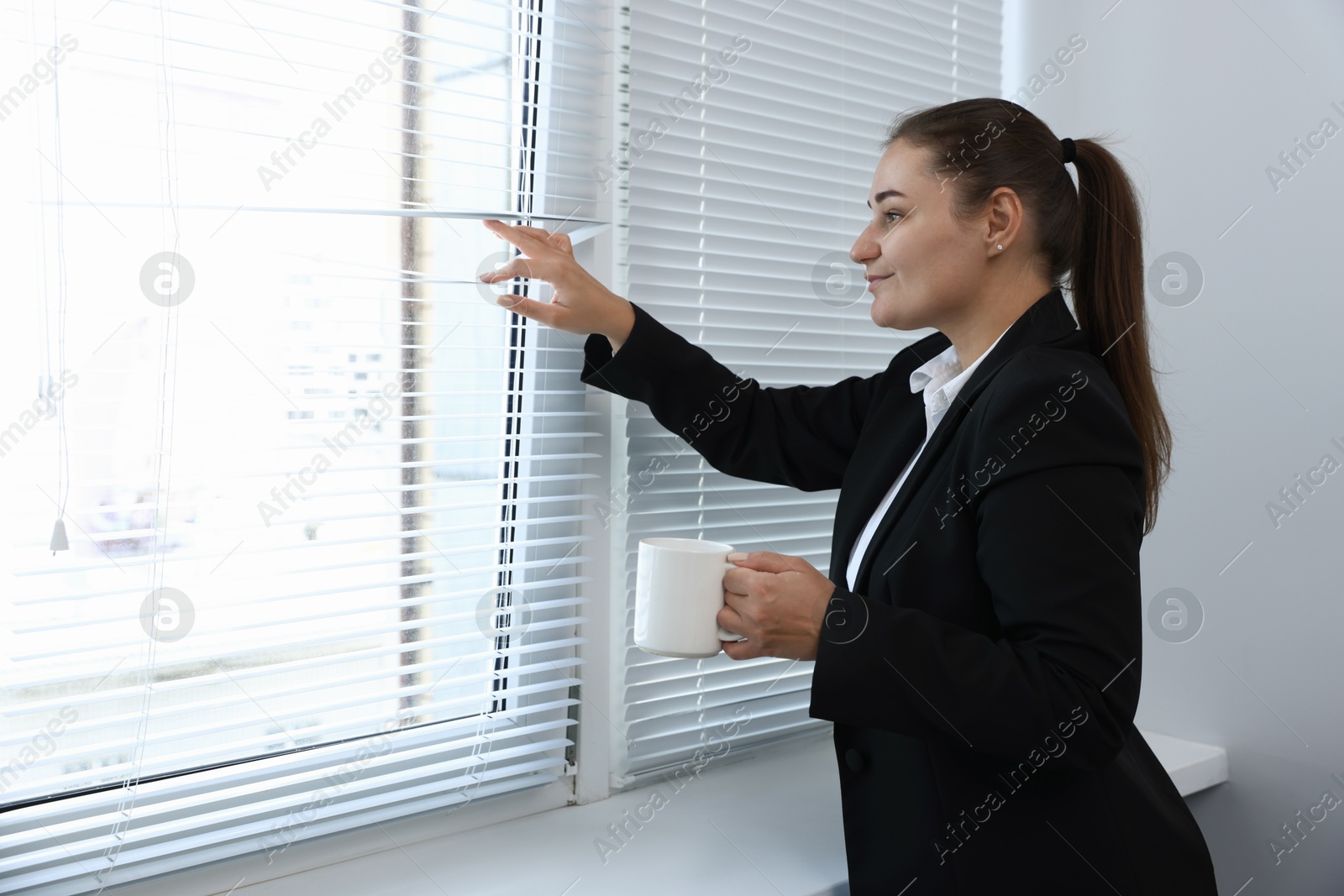 Photo of Curious woman with cup of drink looking through window blinds indoors