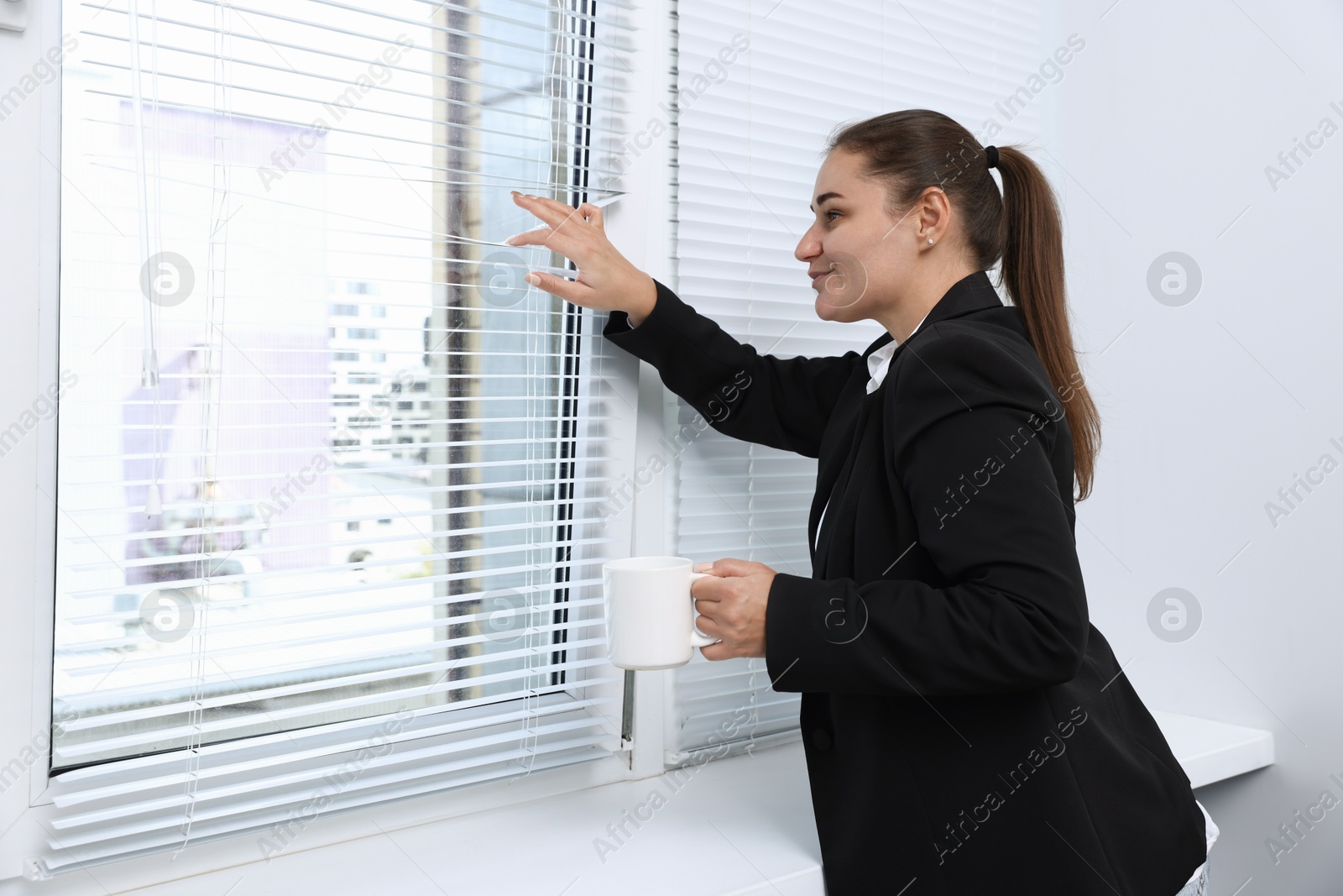 Photo of Curious woman with cup of drink looking through window blinds indoors