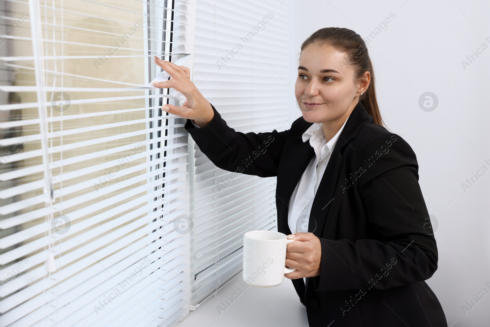 Photo of Curious woman with cup of drink looking through window blinds indoors