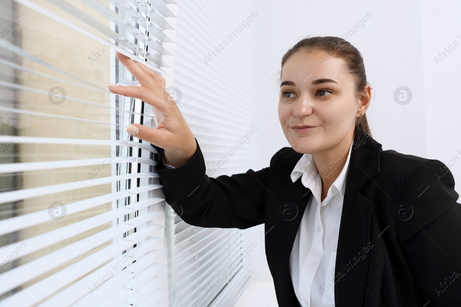 Photo of Curious woman looking through window blinds indoors