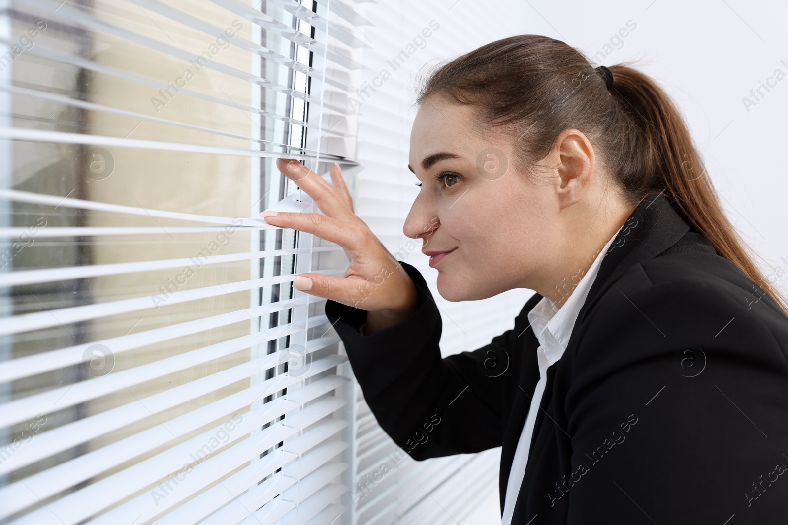 Photo of Curious woman looking through window blinds indoors