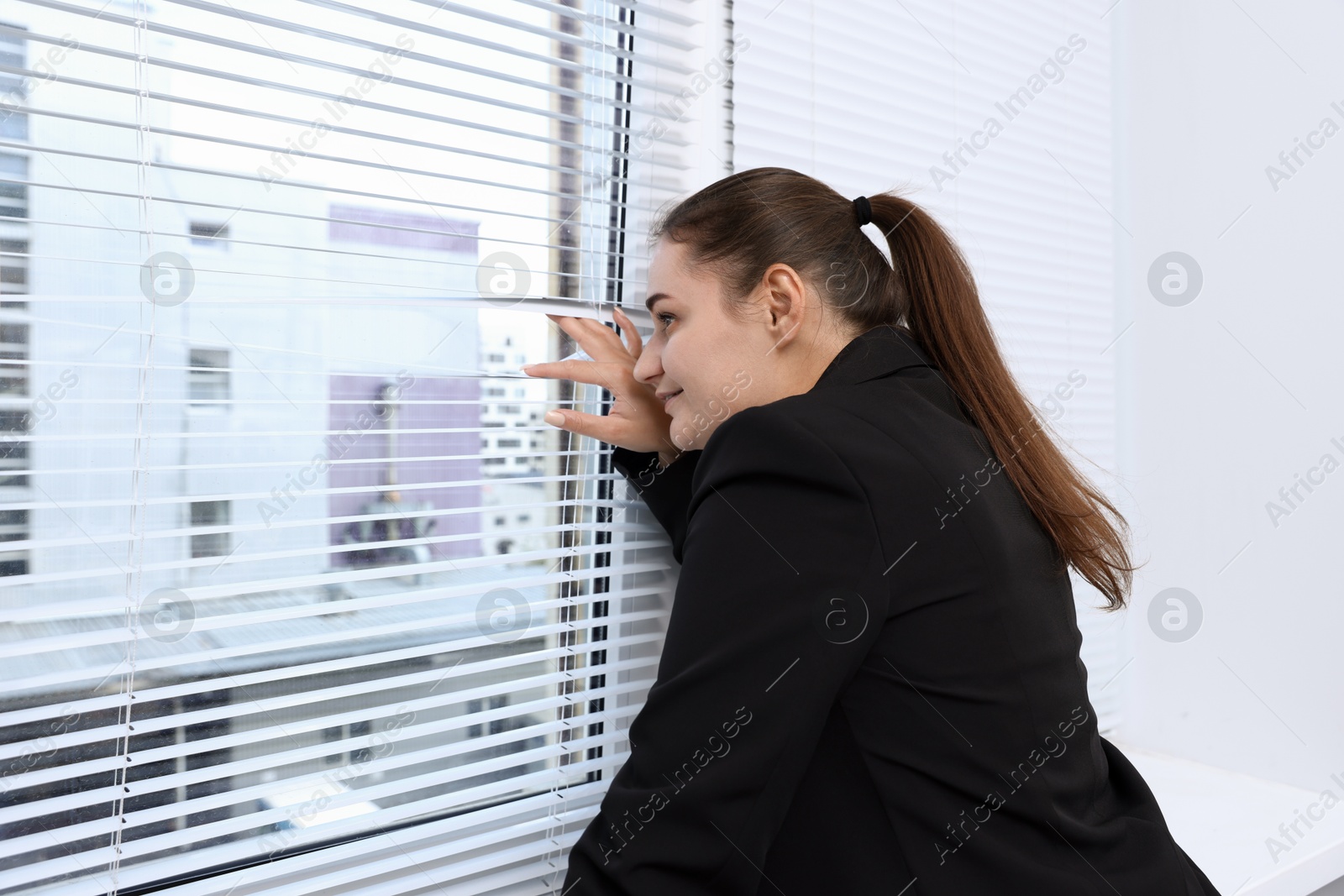 Photo of Curious woman looking through window blinds indoors