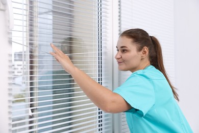 Photo of Curious woman looking through window blinds indoors