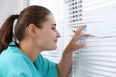Photo of Curious woman looking through window blinds indoors