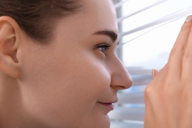 Photo of Woman looking through window blinds indoors, closeup