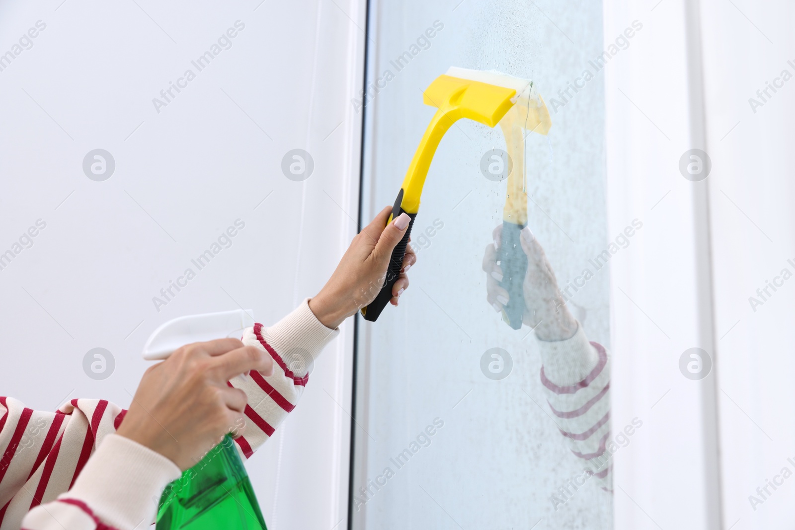 Photo of Woman cleaning window with squeegee and detergent indoors, closeup