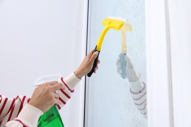Photo of Woman cleaning window with squeegee and detergent indoors, closeup