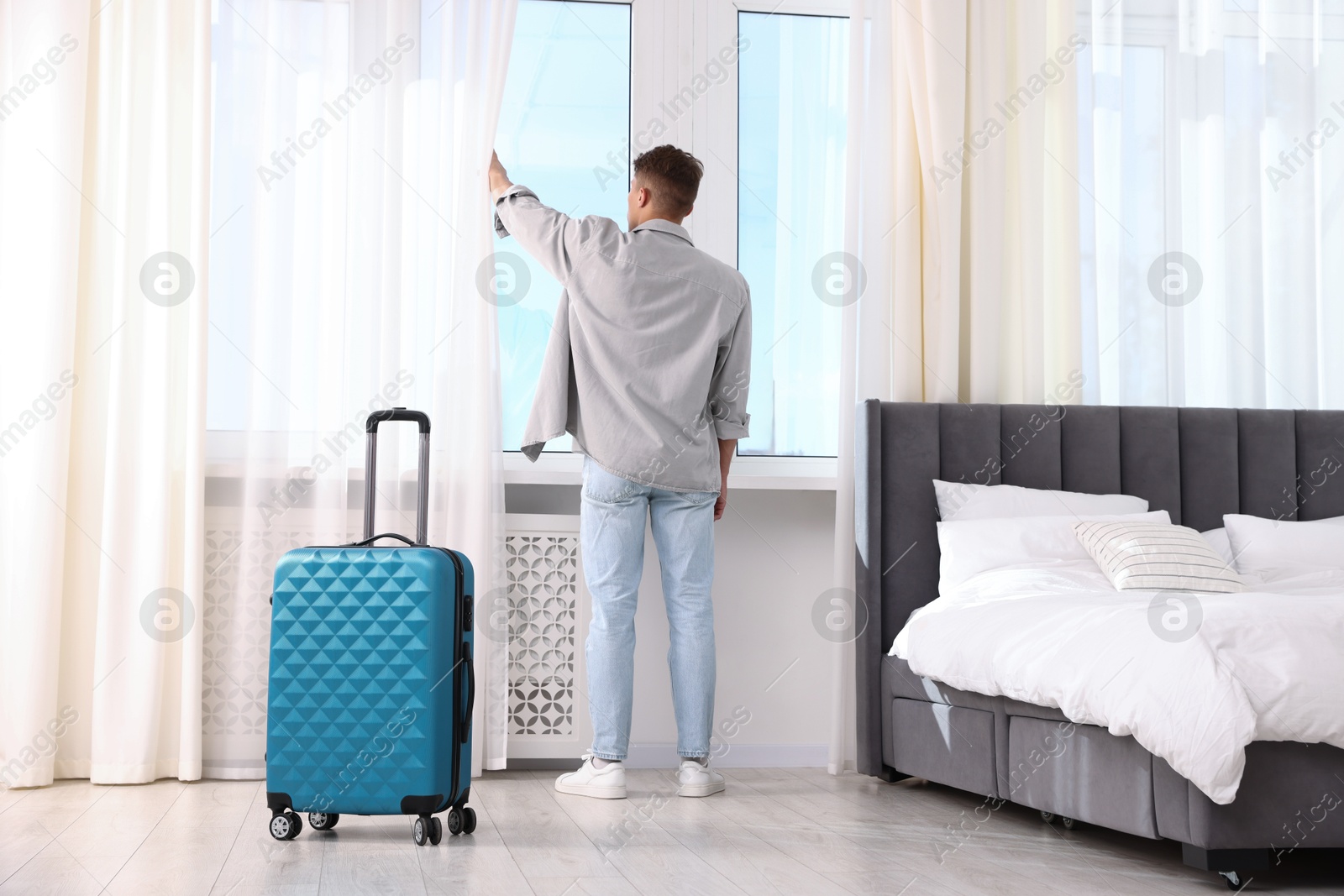 Photo of Man opening curtains and looking out of window indoors, back view