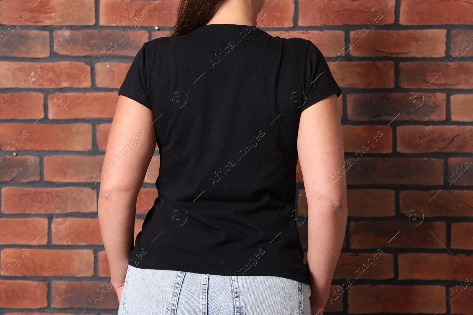 Photo of Young woman wearing stylish black t-shirt near brick wall, back view. Mockup for design
