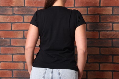 Photo of Young woman wearing stylish black t-shirt near brick wall, back view. Mockup for design