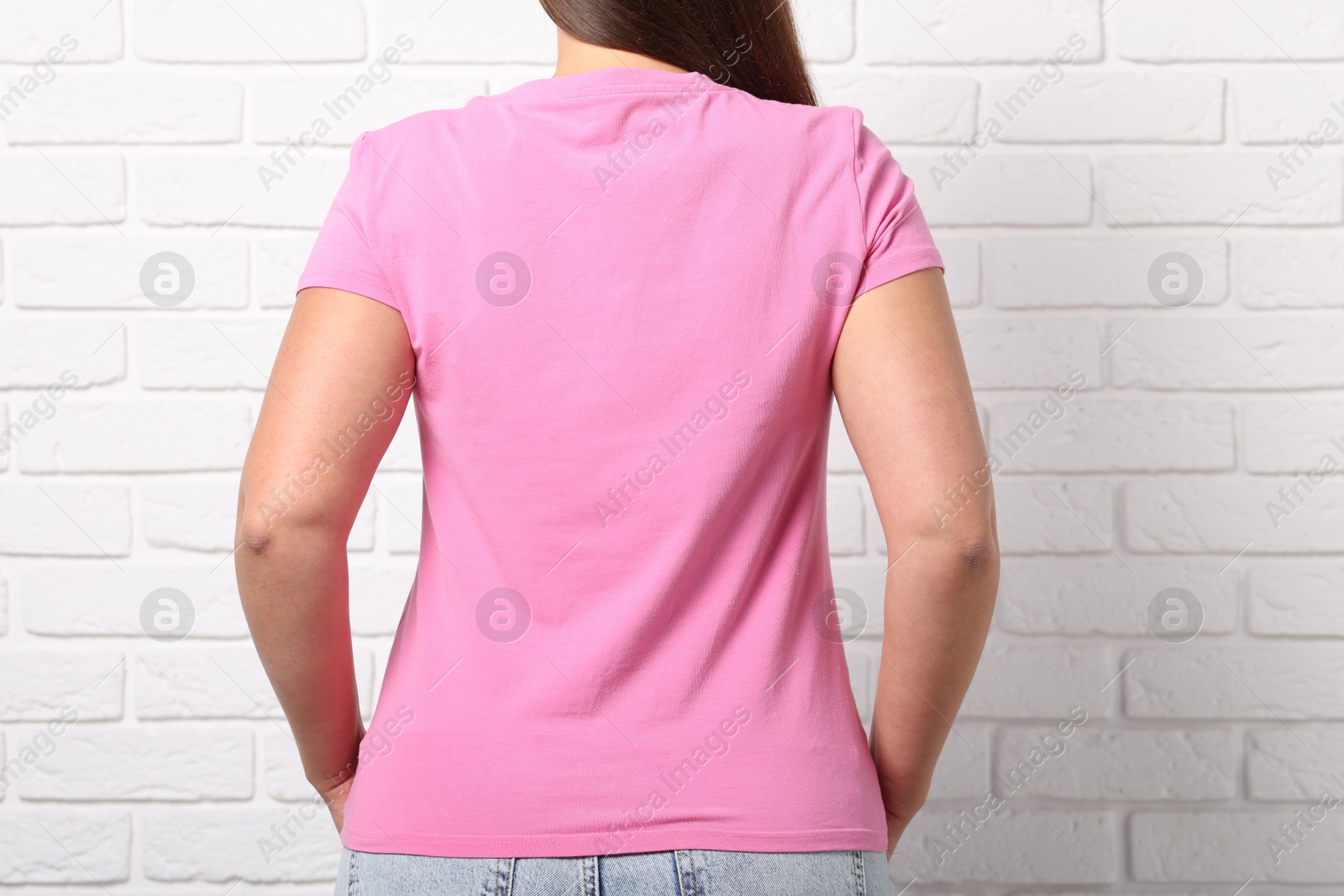 Photo of Young woman wearing stylish pink t-shirt near white brick wall, back view. Mockup for design
