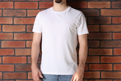Photo of Young man wearing stylish white t-shirt near brick wall, closeup. Mockup for design