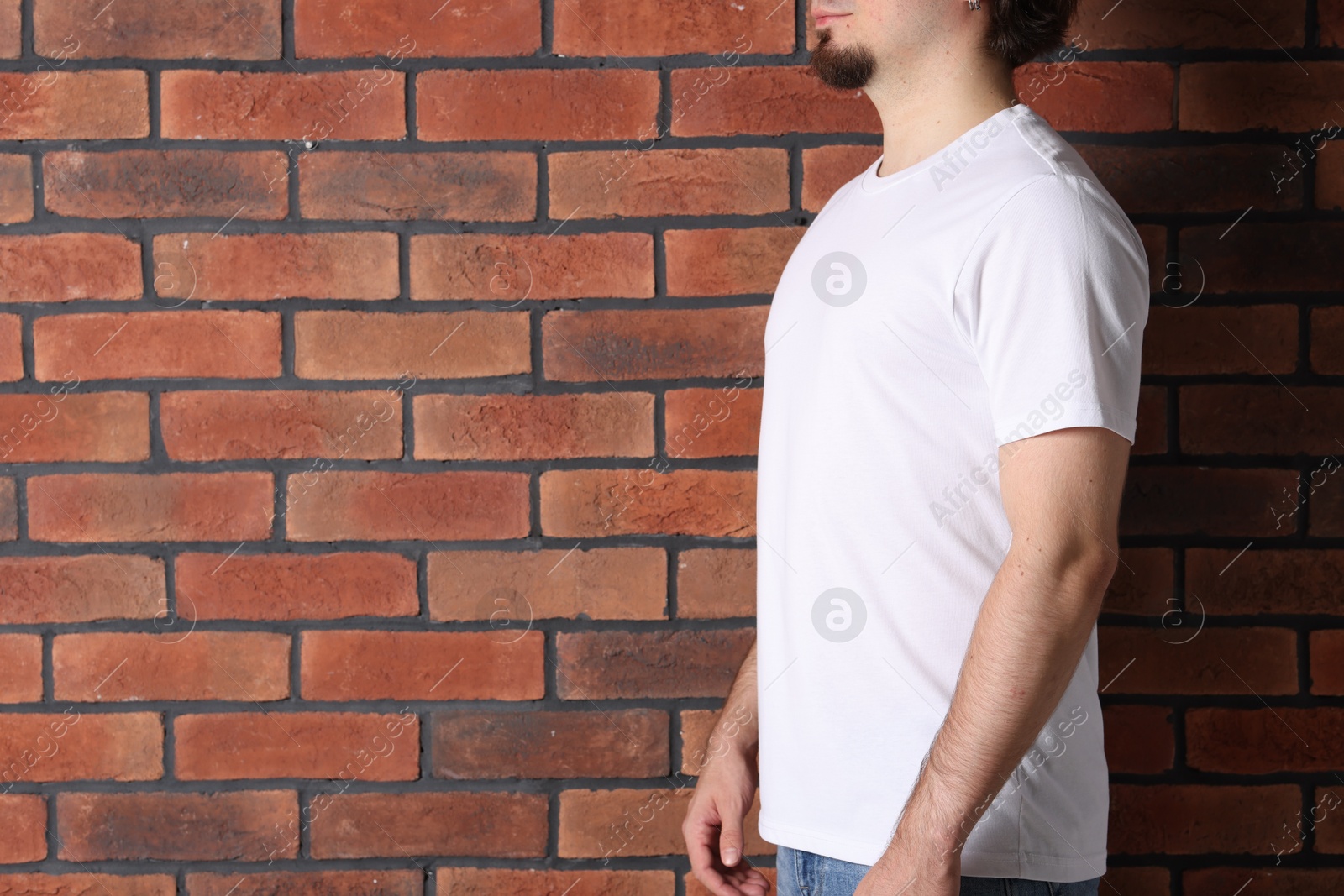 Photo of Young man wearing stylish white t-shirt near brick wall, closeup. Mockup for design