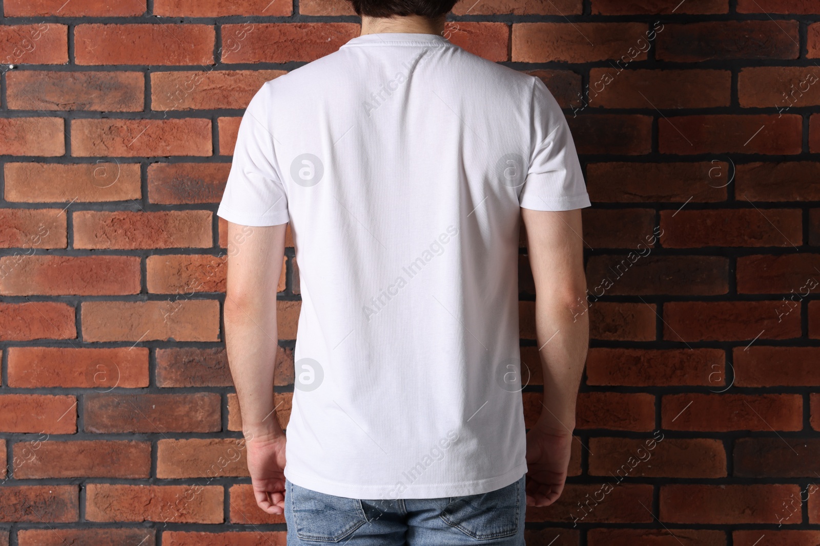 Photo of Young man wearing stylish white t-shirt near brick wall, back view. Mockup for design
