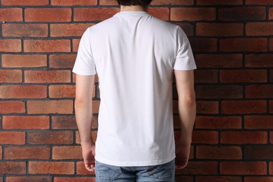 Photo of Young man wearing stylish white t-shirt near brick wall, back view. Mockup for design