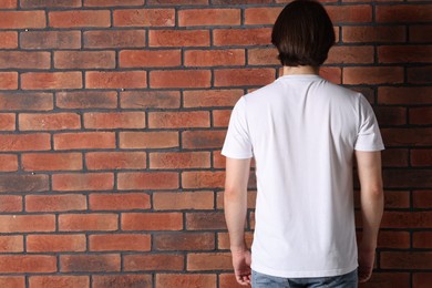 Photo of Young man wearing stylish white t-shirt near brick wall, back view. Mockup for design