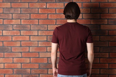 Photo of Young man wearing stylish brown t-shirt near brick wall, back view. Mockup for design