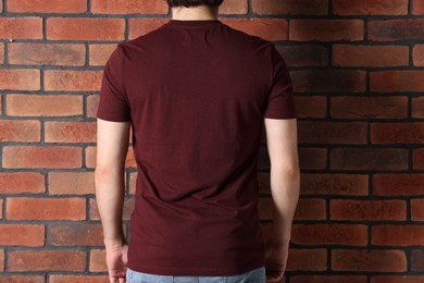 Photo of Young man wearing stylish brown t-shirt near brick wall, back view. Mockup for design