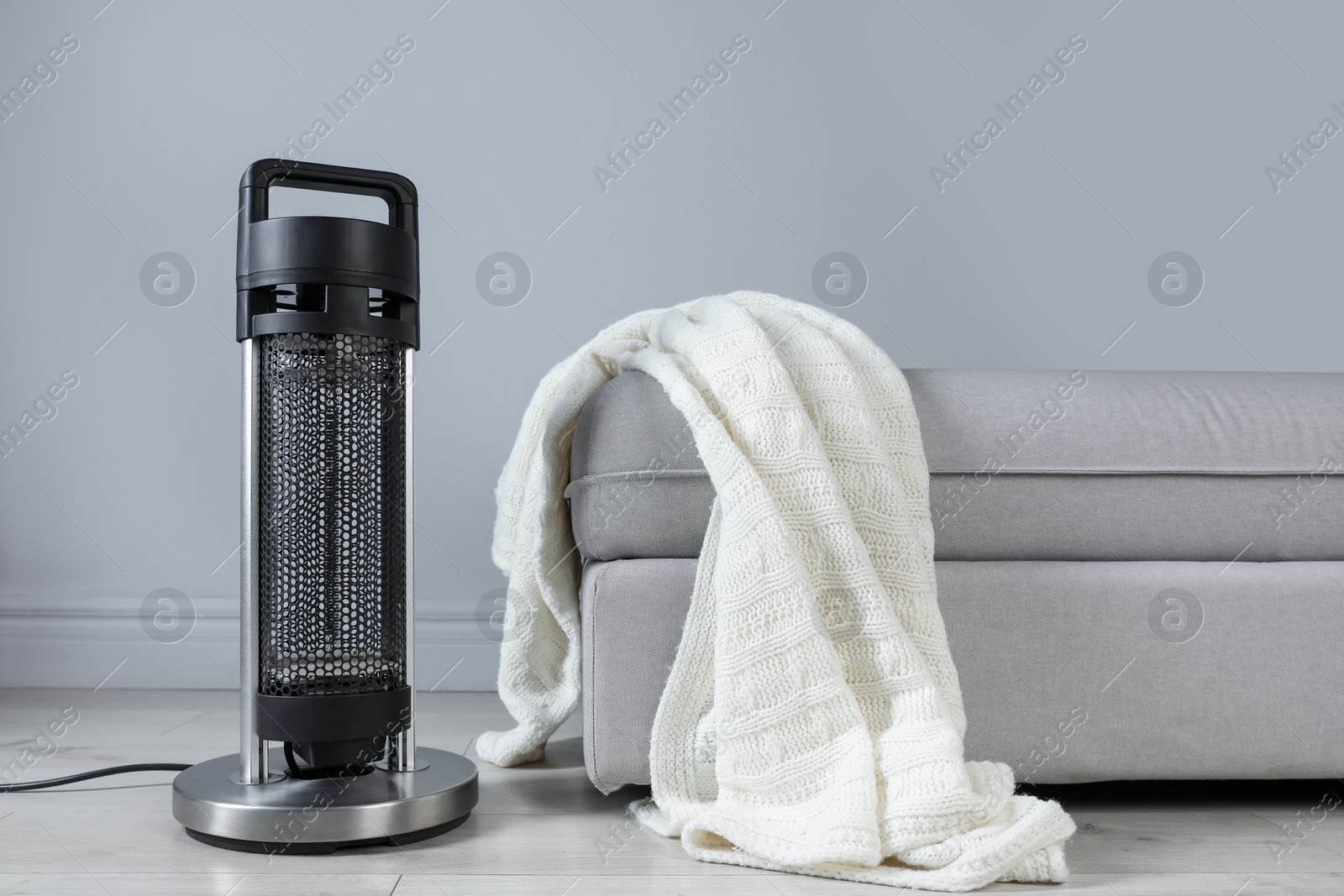 Photo of Electric heater and pouf with blanket on light wooden floor near grey wall indoors