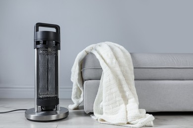 Photo of Electric heater and pouf with blanket on light wooden floor near grey wall indoors