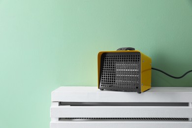 Photo of Electric fan heater on white radiator near green wall indoors. Space for text