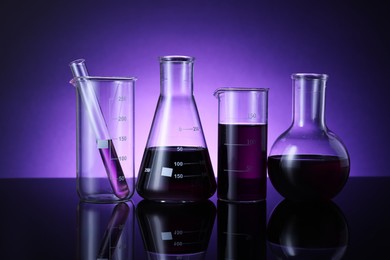 Photo of Different laboratory glassware with color liquid on table against violet background