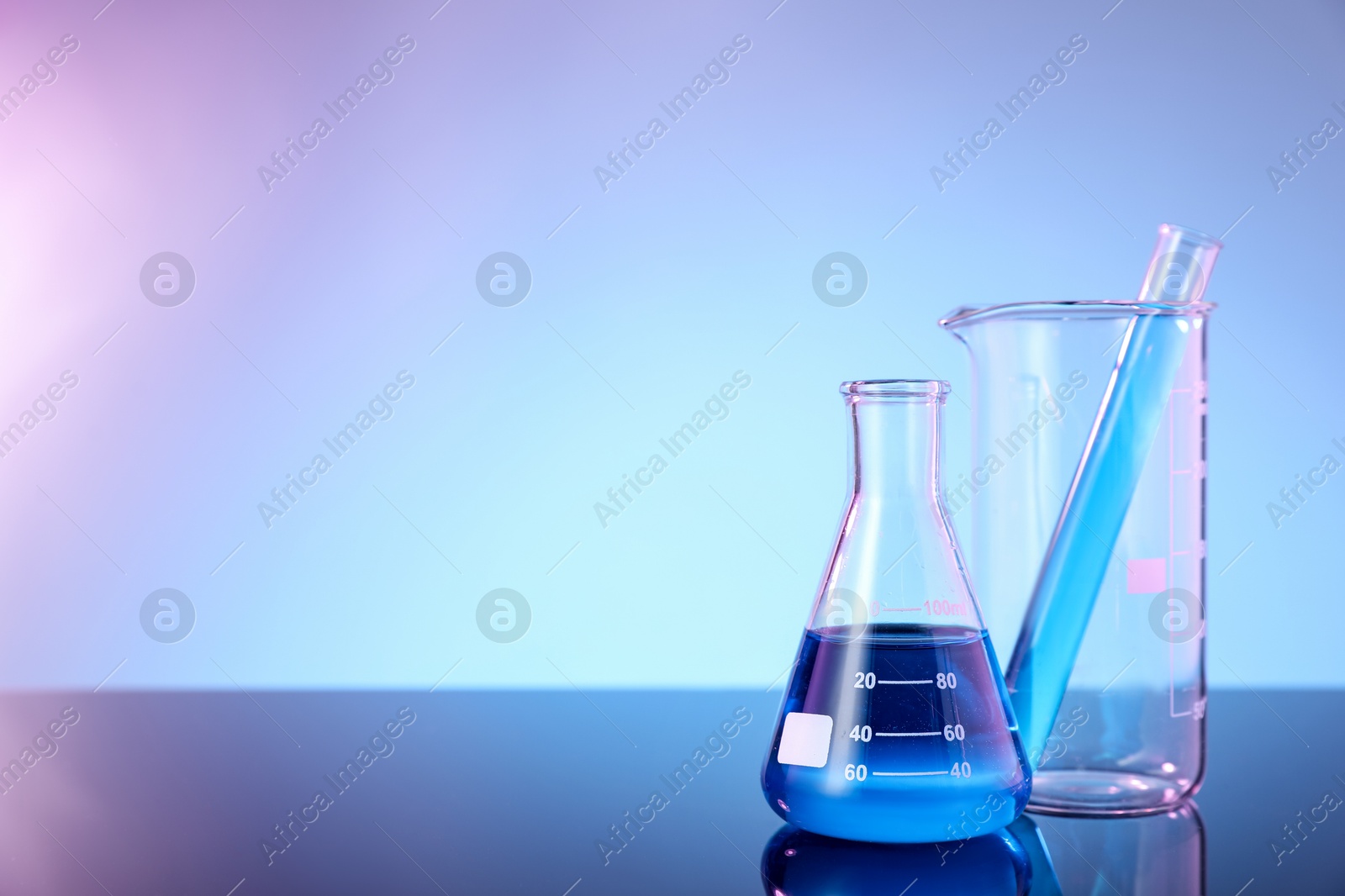Photo of Different glassware with blue liquid on mirror surface against color background, space for text