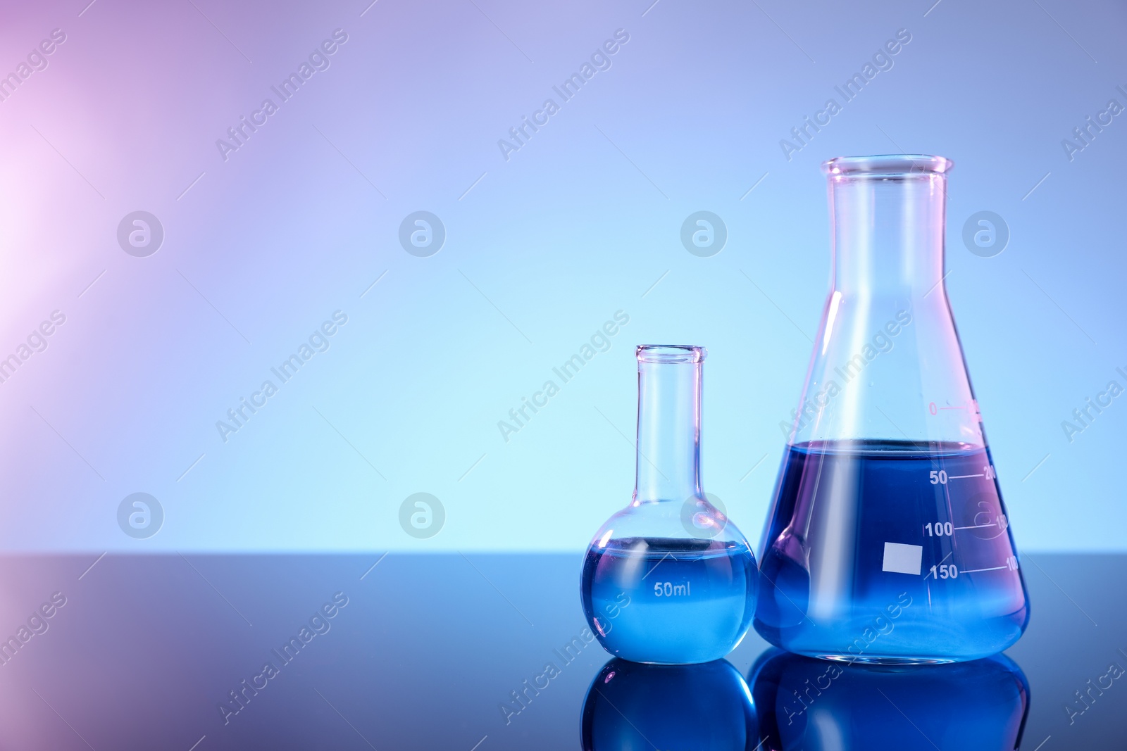Photo of Glass flasks with blue liquid on mirror surface against color background, space for text