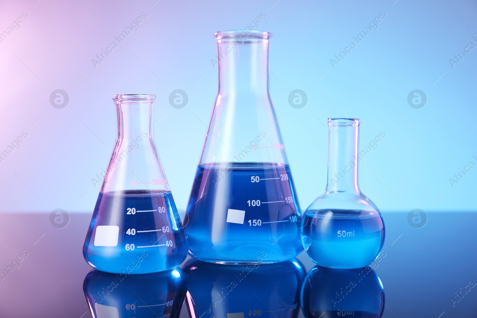 Photo of Glass flasks with blue liquid on mirror surface against color background