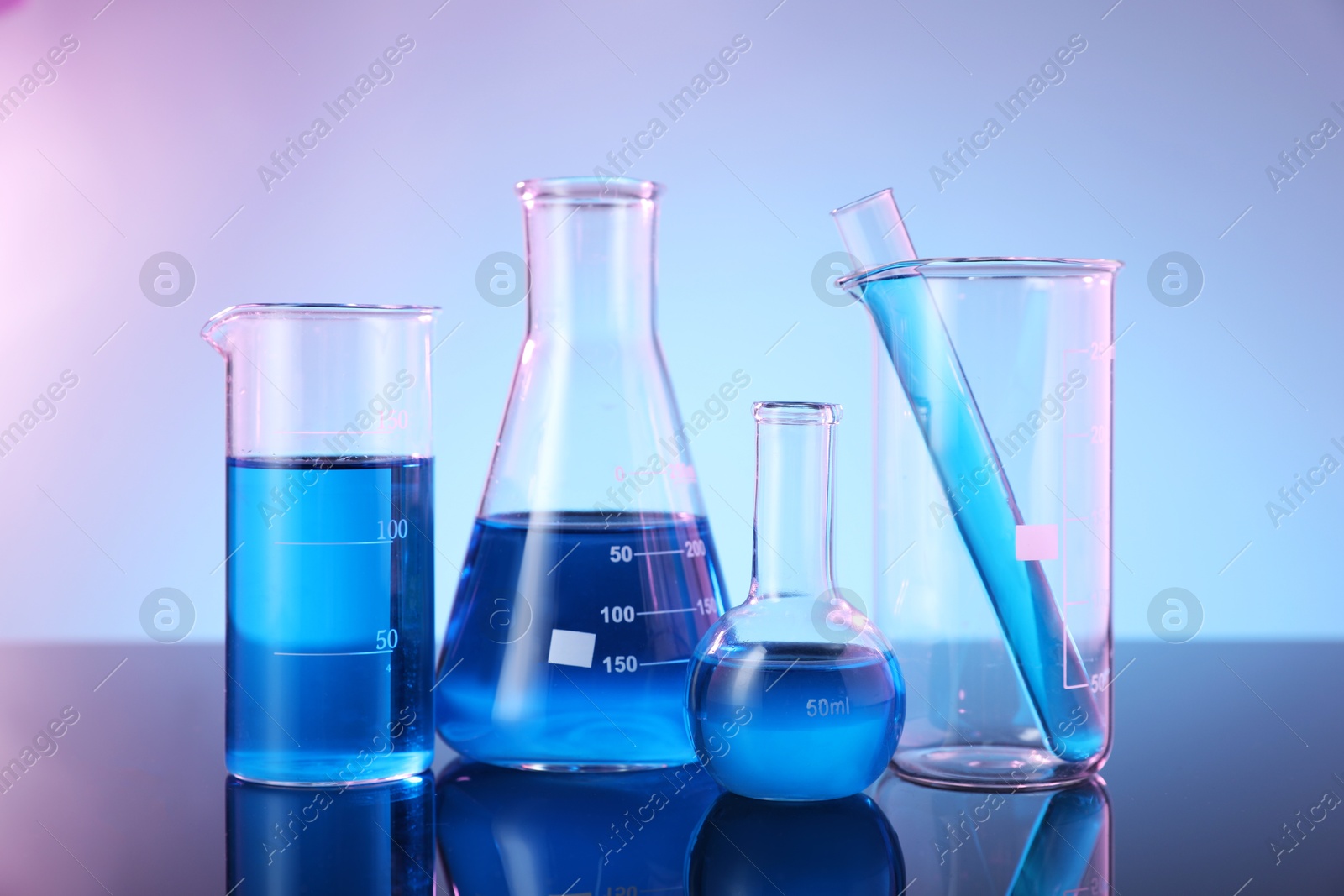 Photo of Different glassware with blue liquid on mirror surface against color background